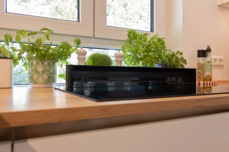 Flächenbündiges Panorama-Kochfeld mit integriertem Dunstabzug vor lichtdurchflutetem Fenster, umgeben von frischen Küchenkräutern auf der Holzarbeitsplatte.