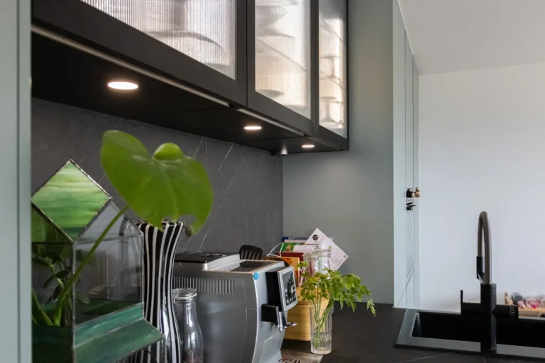 Detailaufnahme einer Küchenzeile mit schwarzer Arbeitsplatte, LED-Unterbauleuchten, Glas-Oberschränken und Kaffeemaschine